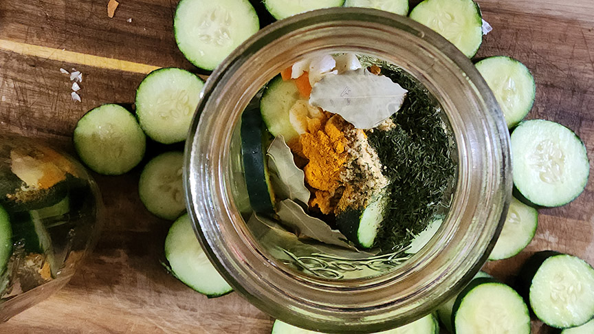 Glass jar filled with pickling herbs and spices surrounded by cucumber slices.