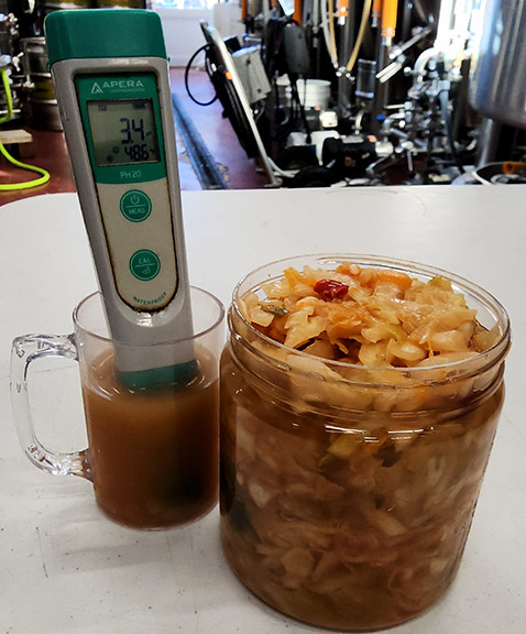 Jar of sauerkraut next to a glass mug filled with liquid with a pH meter.