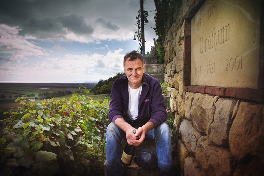 Man sits next to a stone wall near a vineyard holding a bottle of wine.