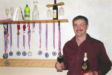 Smiling man holds a bottle and a glass of mead next to a rack of awards and medals.