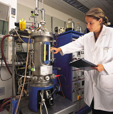 Woman wearing a white lab coat and holding a clipboard inspects equipment used in making yeast.